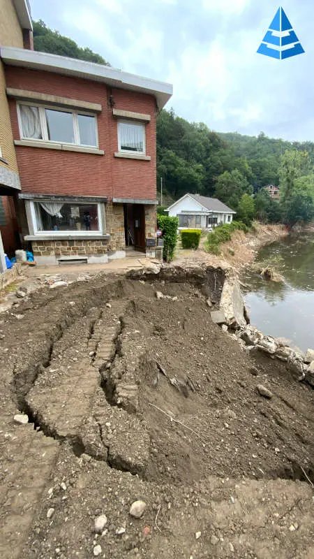 Terrain effondré et fissuré devant une maison en briques, au bord d'une rivière en crue